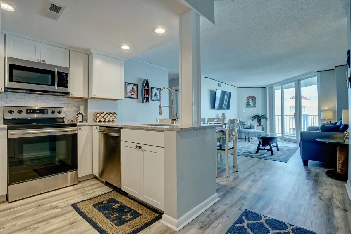 A wide shot of the open-concept living, dining, and kitchen area. The image displays modern kitchen appliances, a comfortable living room seating area, and a dining table, all seamlessly connected with a glimpse of the balcony and ocean beyond.