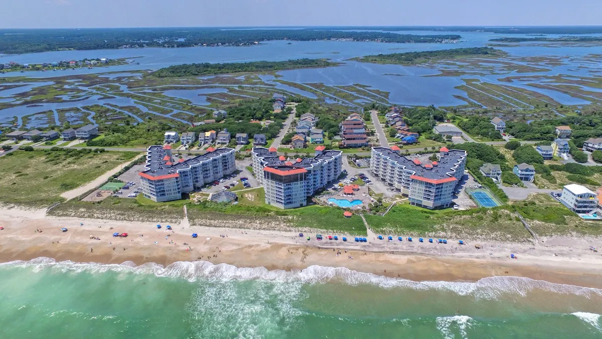 An inviting exterior scene with beachfront buildings, a sparkling swimming pool, sandy beaches, and colorful umbrellas. The lively yet relaxing mood suggests opportunities for sunbathing, socializing, and oceanfront enjoyment just steps away from the property.