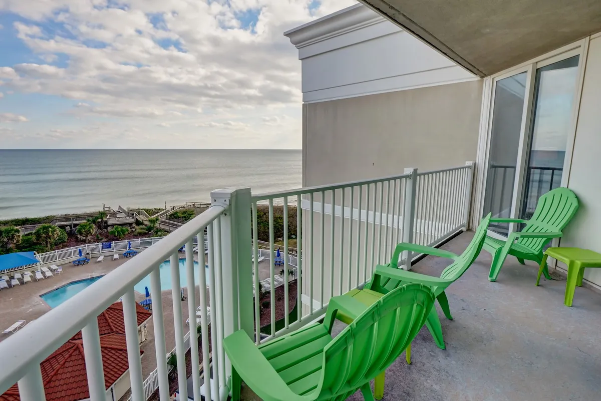 A private balcony scene with green Adirondack chairs inviting guests to sit and enjoy a direct view of the ocean and the pool area below. The mood is tranquil and welcoming, highlighting the outdoor extension of the living space.