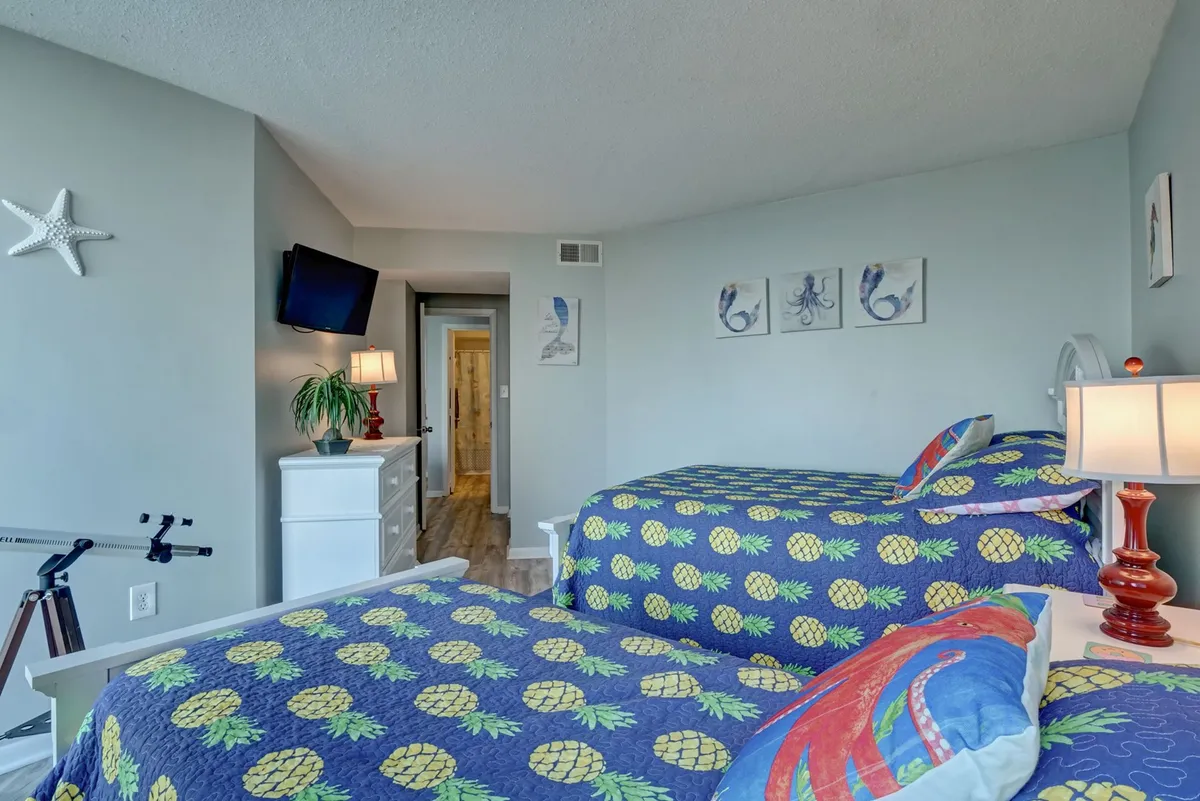 A bright and playful bedroom with two beds adorned with pineapple-patterned covers, a wall-mounted flat-screen TV, decorative sea creature artwork, and a warmly lit bedside lamp. The room exudes a cheerful and relaxed family-friendly vibe.