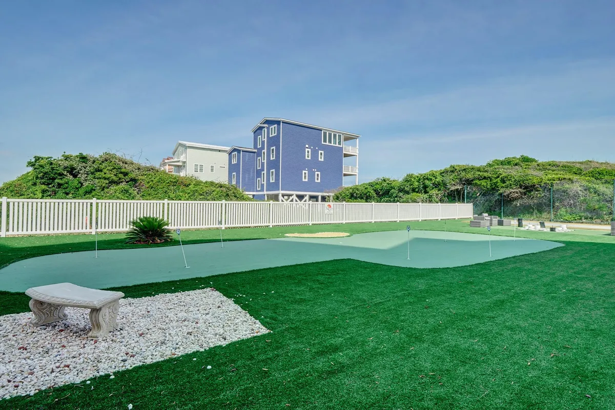 A lush green putting green surrounded by decorative stones and a vibrant blue house, set against a sunny backdrop. The area looks perfect for outdoor activities and small group gatherings, inviting guests to practice golf or simply relax and socialize in the open air.