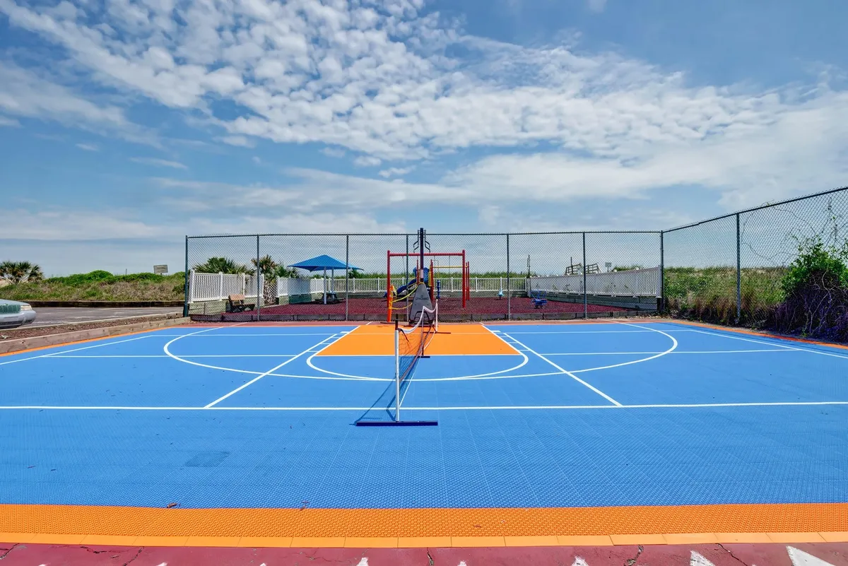 A vibrant outdoor scene featuring a multi-use sports court with both a basketball hoop and a tennis net, alongside modern playground equipment. The space is engineered for active family fun, offering options for pickleball, basketball, and children's play in one area.