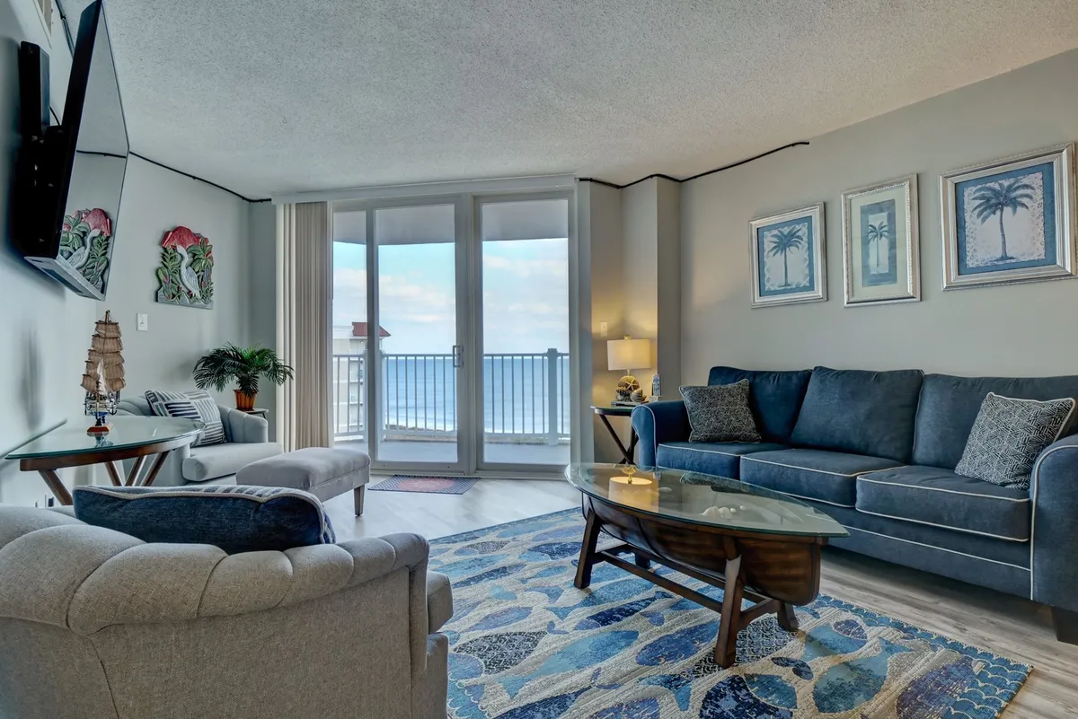 A beautifully appointed living room with large windows framing a stunning ocean view. The space features plush, comfortable seating, a glass coffee table, and tasteful decorative art on the walls, creating an inviting and serene atmosphere perfect for relaxation or socializing.