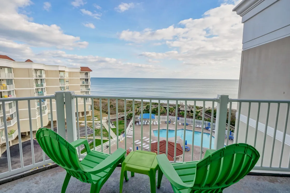 A peaceful view of comfortable green chairs overlooking both the swimming pool and an expansive ocean vista. The mood is tranquil, inviting guests to relax, sunbathe, or simply soak in the views from a prime vantage point.