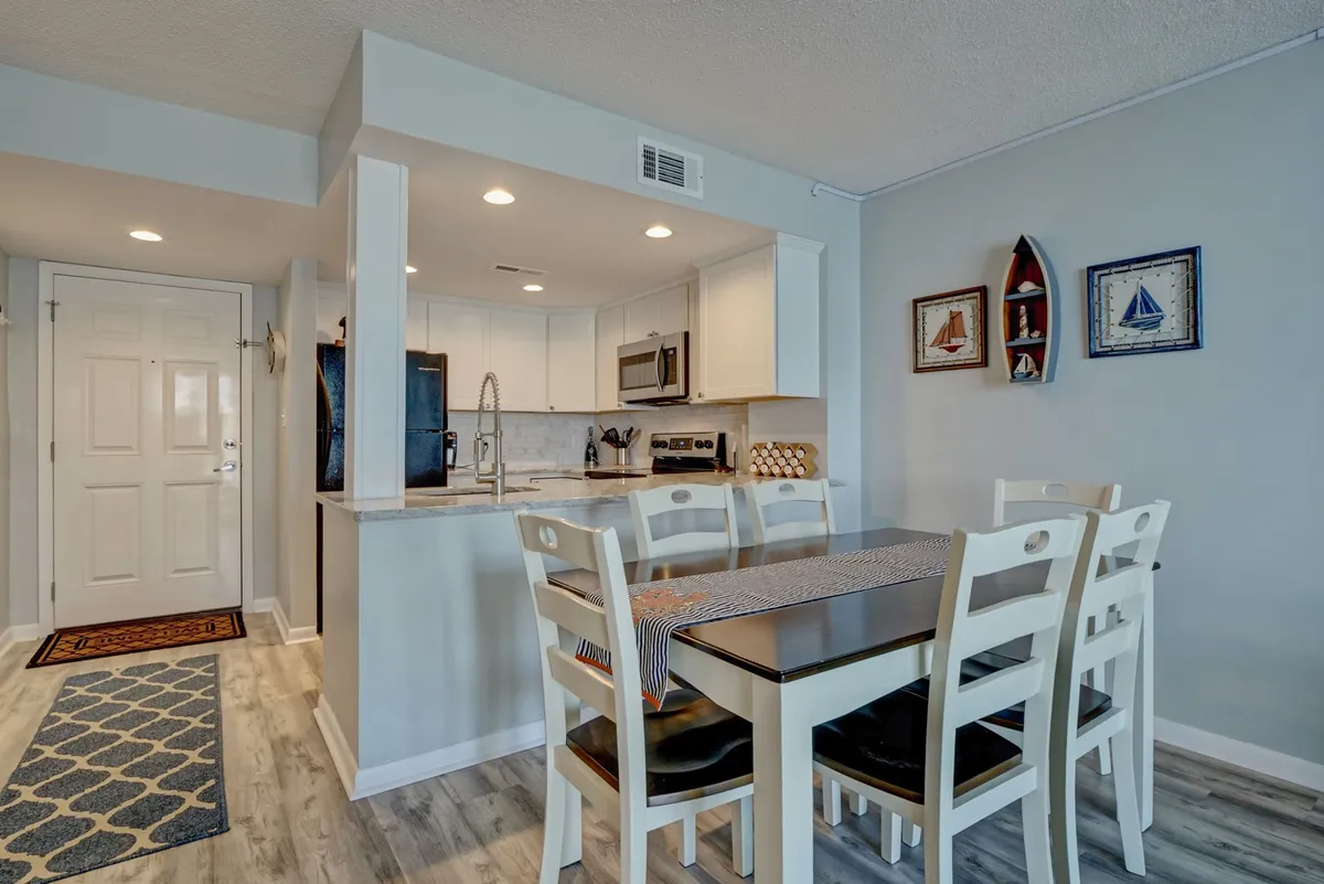 A spacious, light-filled dining area with a table set for a group, adjacent to the kitchen. Decorative wall art and a view toward the entry enhance the ambiance for social meals and gatherings.