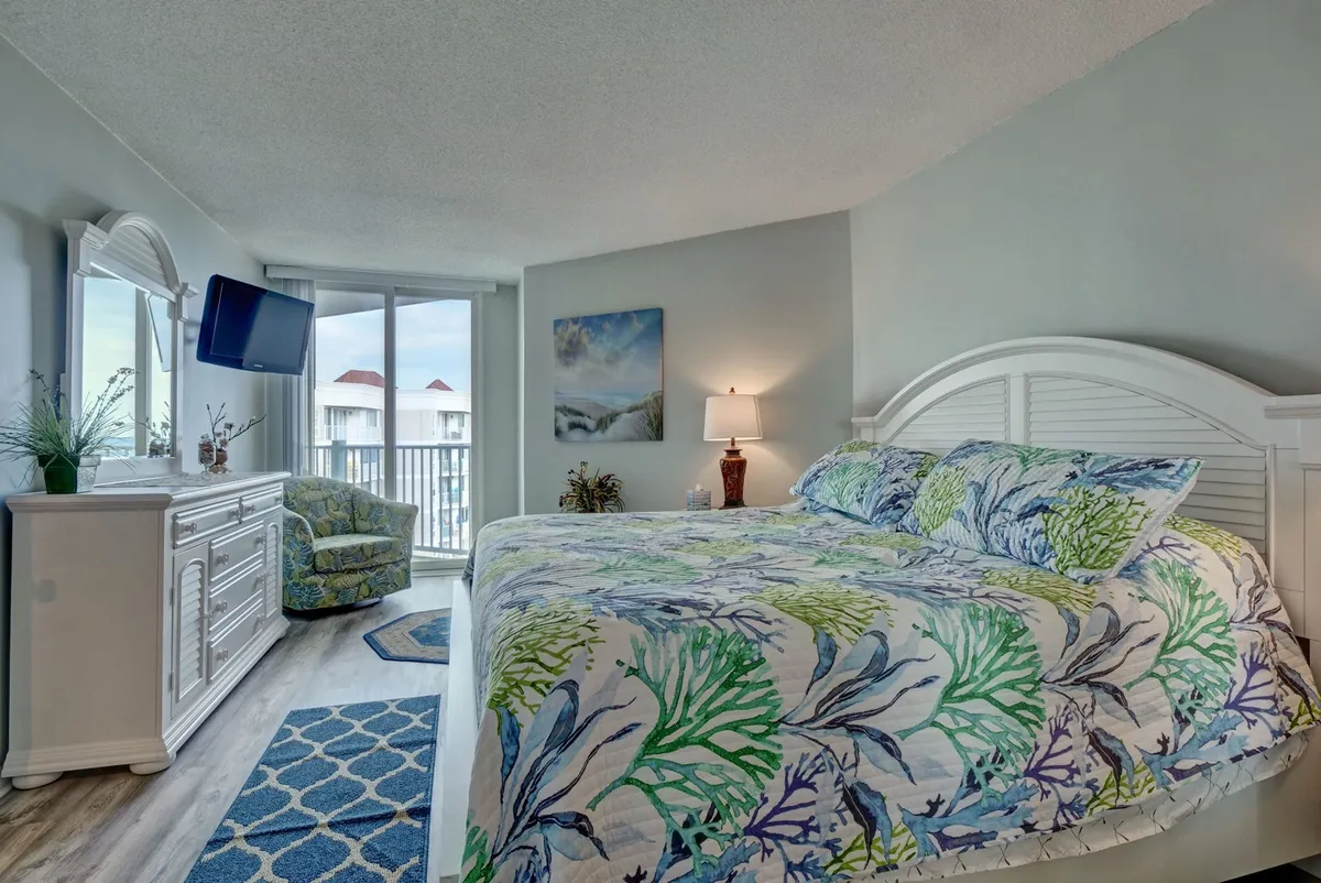 A serene bedroom featuring a plush bed with colorful bedding, a comfortable chair positioned near a window, a wall-mounted television, a decorative mirror, and a softly glowing lamp. Sunlight gently streams into the room, enhancing an inviting and calming atmosphere.