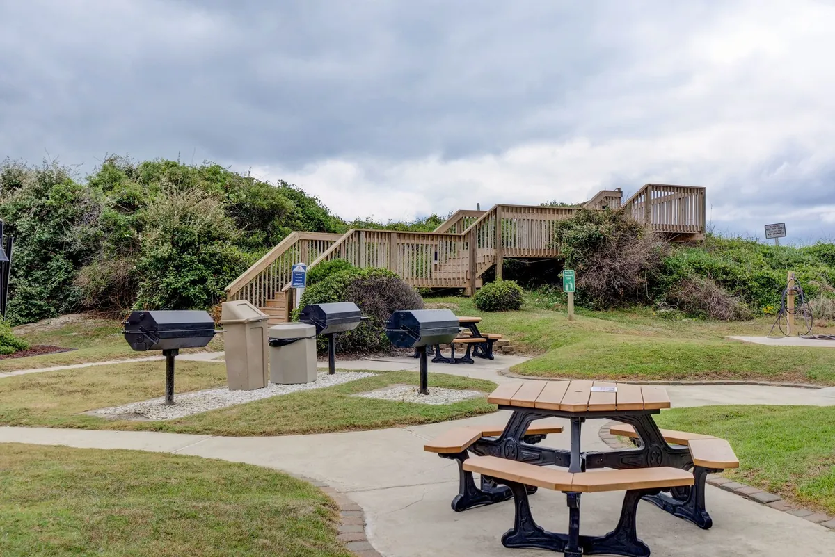 An outdoor grilling and picnic area with several picnic tables, barbecue grills, and a relaxed, communal atmosphere. The space is ideal for dining al fresco and gathering with friends or family.