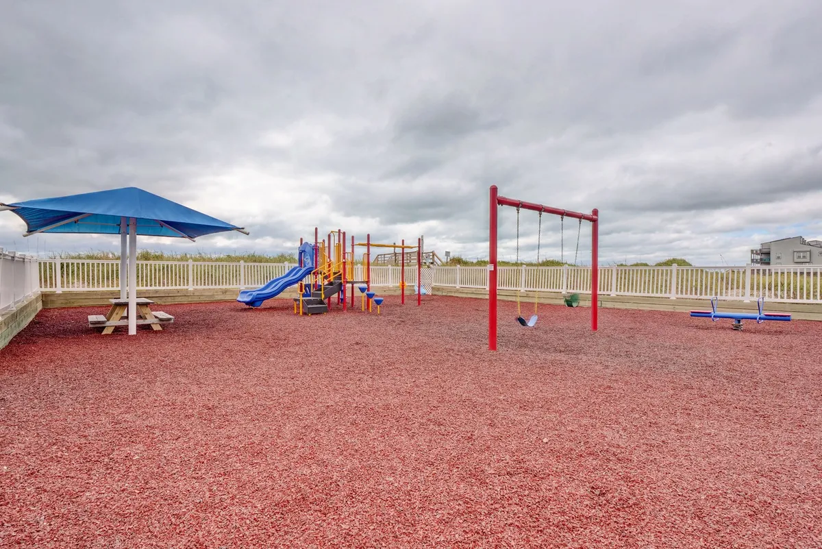 An outdoor playground area with colorful play structures, swings, and a picnic spot shaded by an umbrella, buzzing with joyful activity. The space is designed for families and children to enjoy memorable moments together.